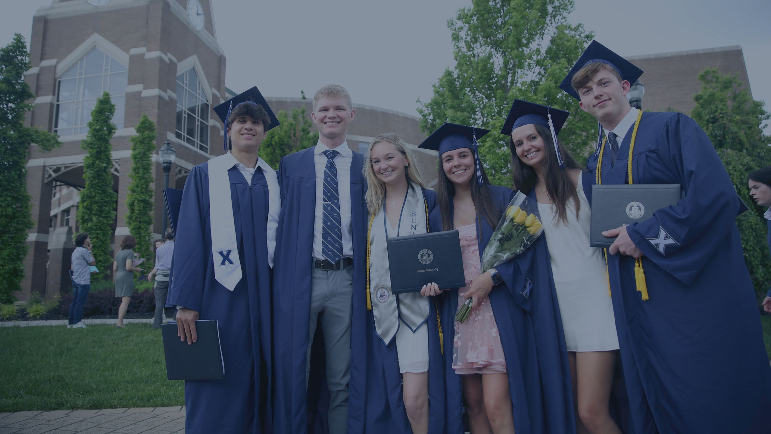 Six Xavier students wearing graduation caps and gowns smile in front of Gallagher Student Center.