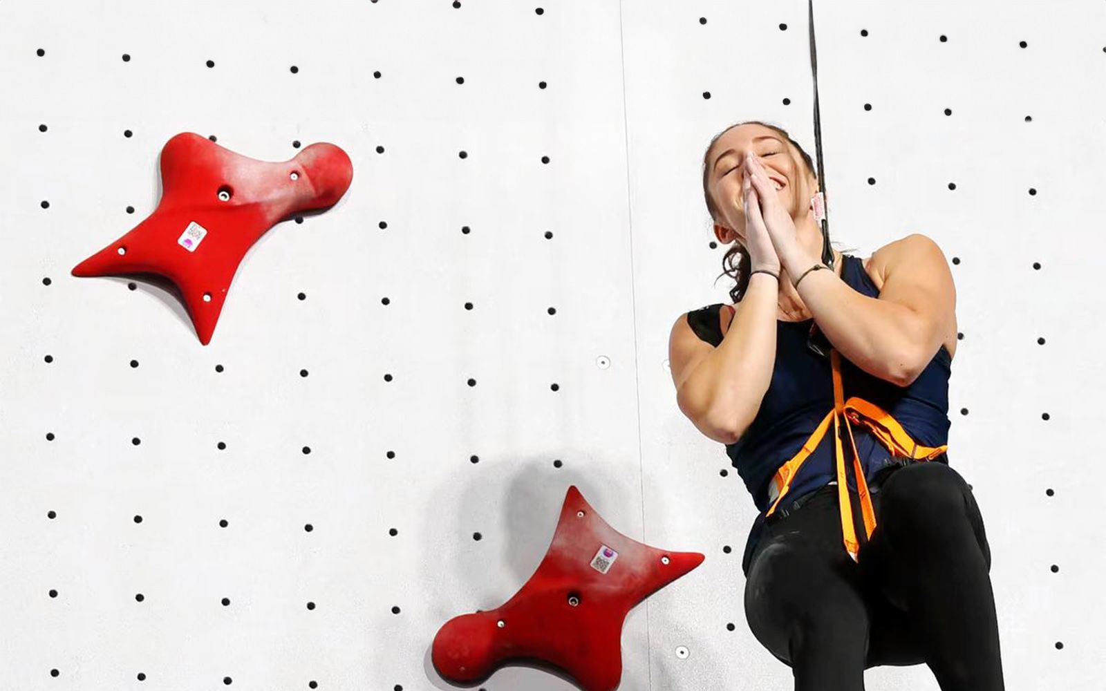 Piper Kelly hanging from side of rock climbing wall as she clasps her hands in gratitude.