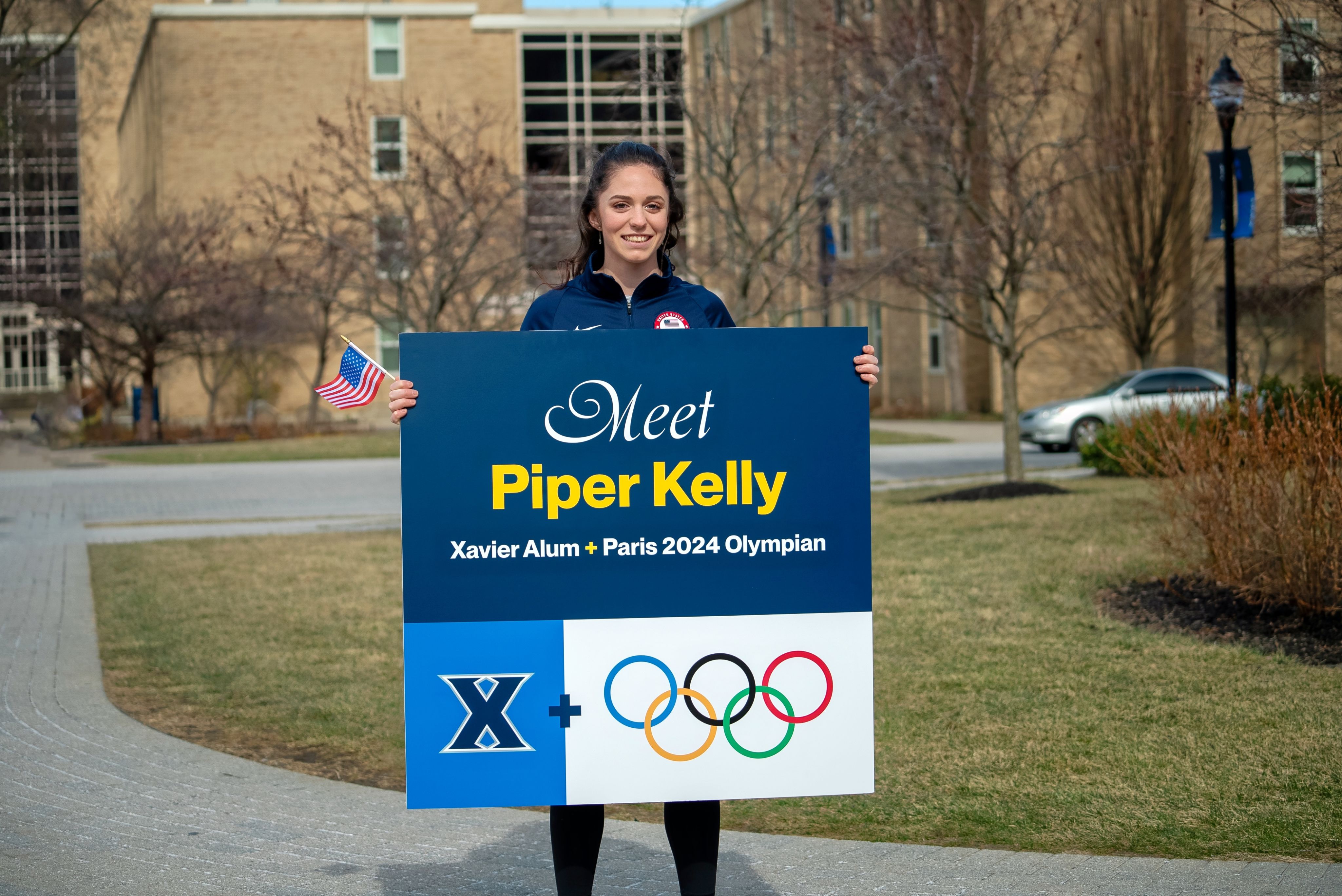 Piper Kelly, holding a sign that says Xavier Alum and Paris 2024 Olympian.