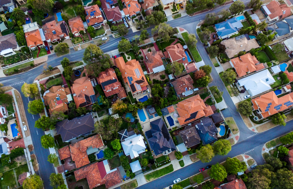 Aerial view of a suburban area near Xavier University, featuring a mix of houses with red, black and white roofing. The streets follow a grid-like, structured layout with neatly arranged homes, lined with large green trees.