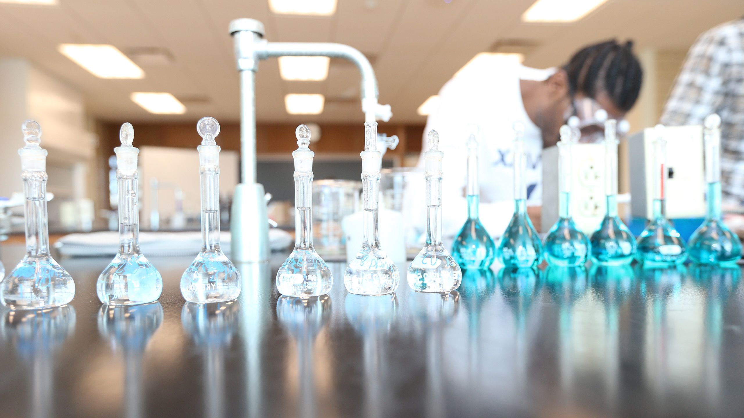 A student in a lab coat students behind several beakers filled with clear and light blue fluid.